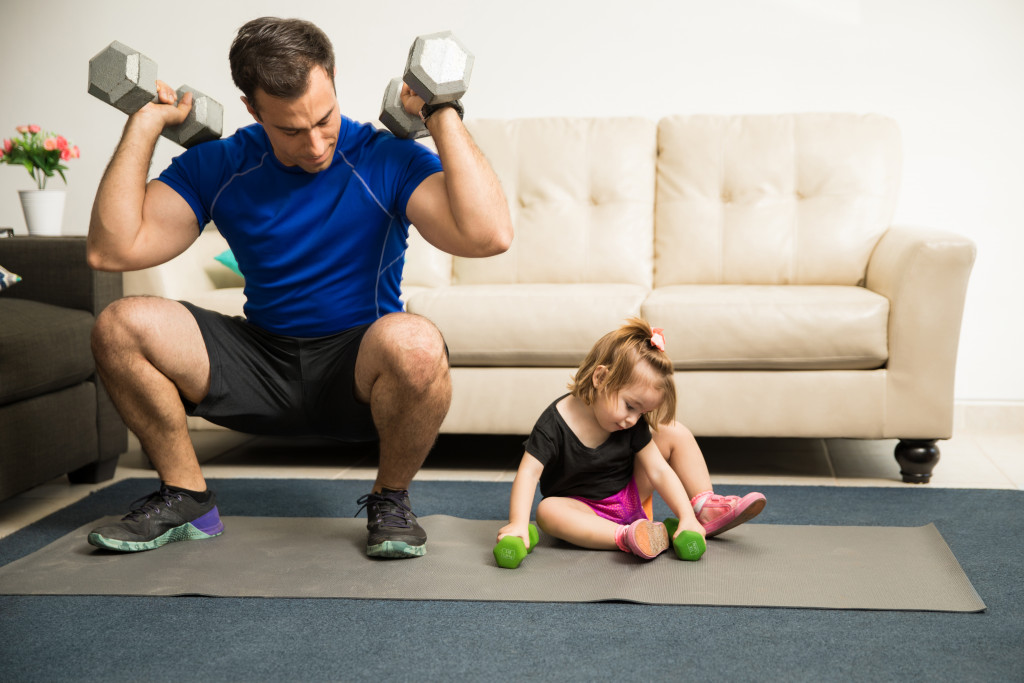 Attractive fitness father showing his strength while his sweet daughter tries to imitate him in the living room